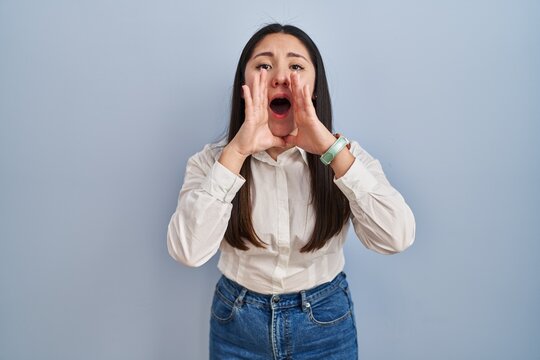Young latin woman standing over blue background shouting angry out loud with hands over mouth