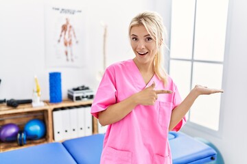 Young caucasian woman working at pain recovery clinic amazed and smiling to the camera while presenting with hand and pointing with finger.