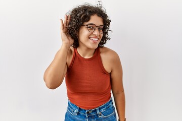 Young hispanic woman wearing glasses standing over isolated background smiling with hand over ear listening an hearing to rumor or gossip. deafness concept.