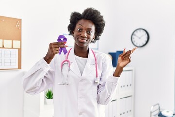 African doctor woman holding purple ribbon awareness at medical clinic smiling happy pointing with...