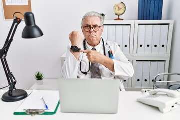 Senior caucasian man wearing doctor uniform and stethoscope at the clinic in hurry pointing to...