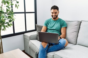 Young handsome man with beard using computer laptop sitting on the sofa at home with a happy and...