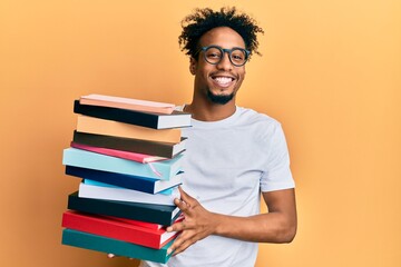 Young african american man with beard holding a pile of books looking positive and happy standing and smiling with a confident smile showing teeth