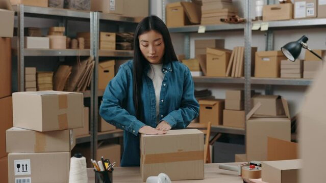Store owner. Cute asian brunette woman putting stickers with client address at the parcel while working at the warehouse. Post service and small business concept.