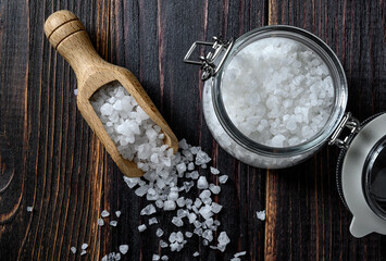 Sea salt in a wooden scoop and glass jar on wooden background. Coarse salt. Top view.
