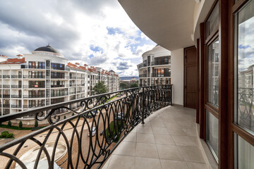 A wide curved balcony of a multi-storey building with metal black wrought iron railings with patterns. The balcony offers a view of the houses of the residential complex, mountains and clouds