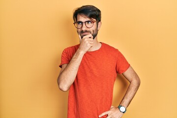 Young hispanic man wearing casual clothes and glasses with hand on chin thinking about question,...