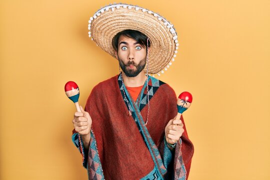 Young hispanic man wearing mexican hat holding maracas making fish face with mouth and squinting eyes, crazy and comical.