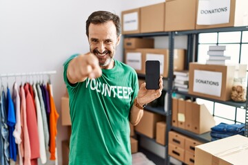 Middle age man with beard wearing volunteer t shirt holding smartphone pointing to you and the...