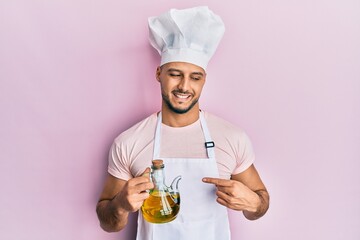 Young arab man wearing professional cook uniform holding olive oil can smiling happy pointing with...