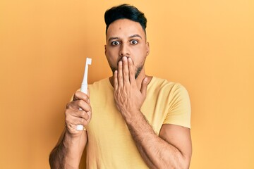 Young arab man holding electric toothbrush covering mouth with hand, shocked and afraid for...