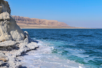 View of the mountains across the waters of the Dead Sea from the shore covered with salt formations.