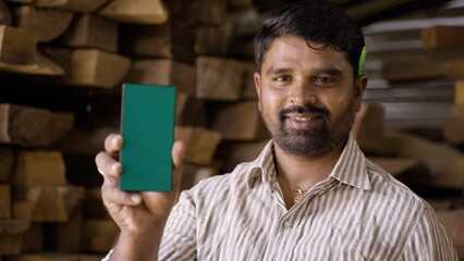 Rack focus shot, happy smiling Indian artisan with green screen mobile phone looking at camera at carpentry shop - conept of online booking of woodworking service, advertising and blue collar jobs