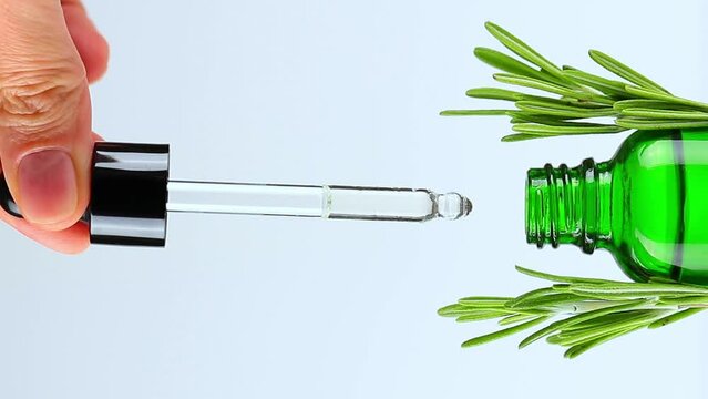 Close-up of female hand lifts dropper lid, oily drops fall from cosmetic pipette. Green dropper bottle with natural cosmetic oil. Fresh herbs rosemary on each side of bottle. Vertical format video.