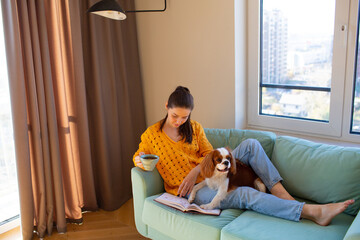 Young woman sitting at home on sofa with Cavalier King Charles Spaniel dog and read a book.