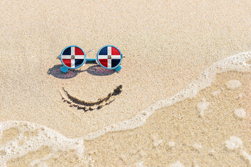 Fototapeta na wymiar A painted smile on the sand and sunglasses with the flag of the Dominican Republic. The concept of a positive and successful holiday in the resort of the Dominican Republic.