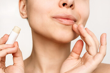 Cropped shot of a young caucasian woman touching her lips with the finger and holding a hygienic lipstick in her hand isolated on a white background. Moisturizing chapstick for dry lips. Close up - obrazy, fototapety, plakaty