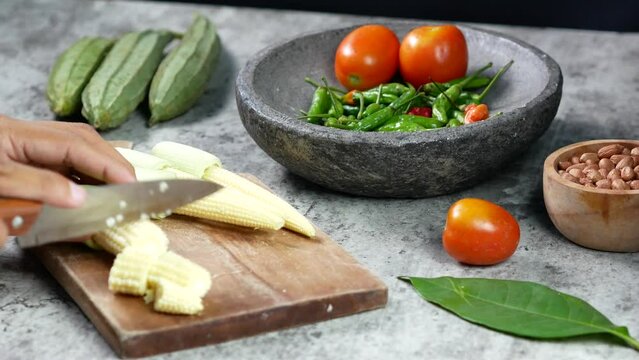 hand slicing baby corn with knife on wooden cutting board.