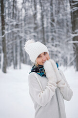 Portrait of young Caucasian woman dressed up white sweater, knitted hat with pompom, fluffy mittens, green scarf and dress walking alone in park or forest on snowy winter day. Ice skating in winter