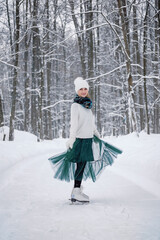 Portrait of young Caucasian woman dressed up white sweater, knitted hat with pompom, fluffy mittens, green scarf and dress ice skating alone in park or forest on snowy winter day. Winter activities.