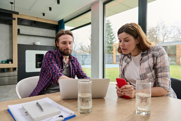 Beautiful surprised pregnant woman holding a smartphone in her hands and looking at the laptop monitor sitting near her husband who showing her an information on the screen and smiling cheerfully