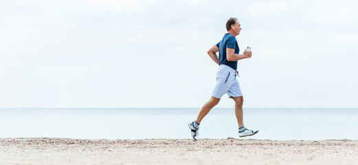 Elderly man runs early morning by sea beach. Active senior man is jogging and listening music. Healthy retirement lifestyle.