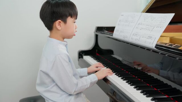 Zoom out of Asia boy playing grand piano in the classroom. Kid wearing casual shirt. Child looking at paper and having fun and relax during class. Education, skill and learning concept