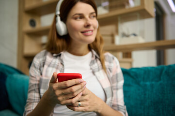 Soft focus on the hands with smartphone of a blurred Caucasian pregnant beautiful woman dreamily looking aside and smiling with cheerful toothy smile, listening to the music in wireless headphones