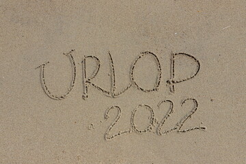 The inscription urlop 2022 in Polish, vacation 2022, on the sand by the water and the rising wave, seashore beach vacation by the sea