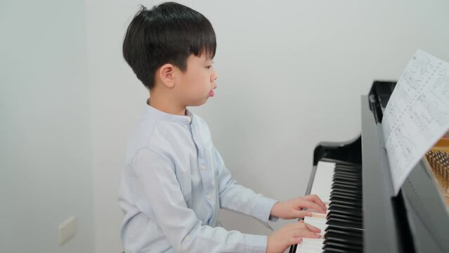 Asia boy wearing shirt and playing grand piano in the classroom at school. Child looking at paper note and having fun during class. Education, skill and learning concept. Musician piano practice