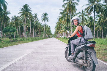 Tropical travel and transport. Young beautiful woman in helmet riding scooter on the road with palm...