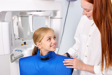 Professional Redhead Female dental specialist is going to examine an x-ray of child girl in modern dental clinic by professional specialist. little caucasian kid has dentist checkup. Portrait