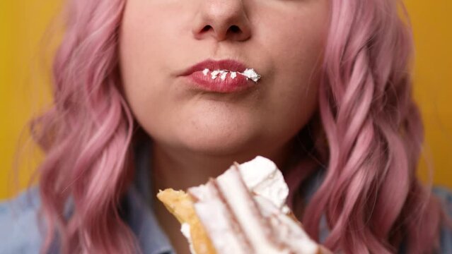 Closeup of attractive cheerful hungry famale girl with pink hair hairstyle enjoying biting tasty sweet cream cupcake isolated over yellow background