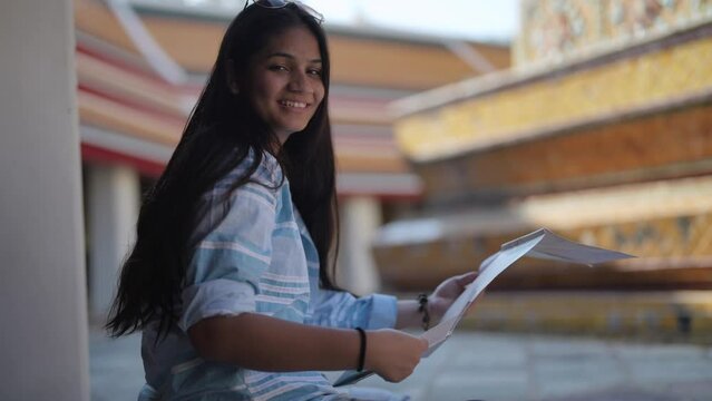 Asian woman use generic local map while sitting on pedestrian terrace. Young teen backpacker travel to sightseeing a landmark temple in Bangkok. Thailand.