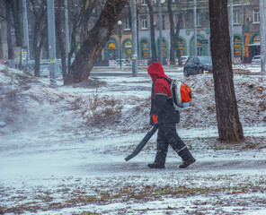 Seasonal work of city utilities in the park. A worker with a motorized backpack blower blows snow...