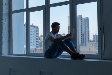 African American man in glasses and a white t-shirt uses a smartphone while sitting by the window.