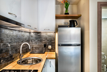 Modern kitchenette corner in grey colors. Equipped with big fridge, electrical kettle, toaster, water sink.