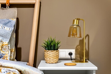 Bedside table with greenplant in straw pot and stylish glass desk lamp in gold, brass and caramel color. Interior design concept.  Selective focus.