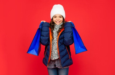 happy kid in puffer jacket and hat. teen girl after shopping on red background.