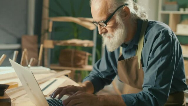 A Focused Carpenter Develops Layouts from Wood, Models 3D Sketches Using a Laptop in a Home Workshop. Garage Hobby, Profession, Joinery and New Computer Technology.