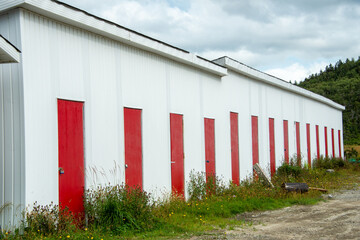 Fototapeta na wymiar The exterior of a white concrete commercial building with a flat roof, multiple single vibrant red metal doors. There's grass in front of the storage unit, and a hill and clouds in the background.