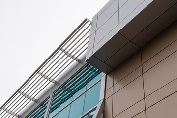 The upward view of a modern commercial building with beige and grey metal composite panels, blue glass windows, and a slated grate on the edge of the roof. The sky is cloudy and white in color. - Powered by Adobe