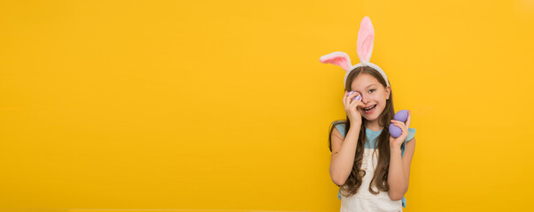 Happy little girl with long dark hair in a headband with hare ears holds easter purple eggs in her hands and smiles while stroking at the camera, easter banner with a smiling girl on a yellow