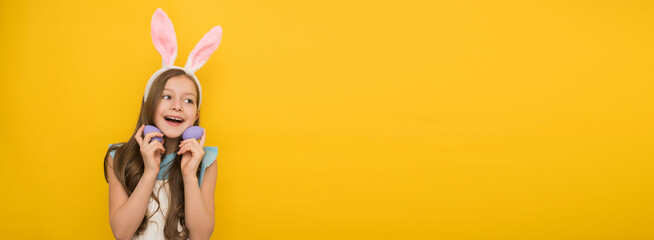 Happy little girl with long dark hair in a headband with hare ears holds easter purple eggs in her hands and smiles while stroking at the camera, easter banner with a smiling girl on a yellow