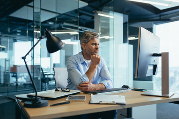 A gray-haired man thinks about the tasks, a businessman with a beard works in a modern office at the computer,