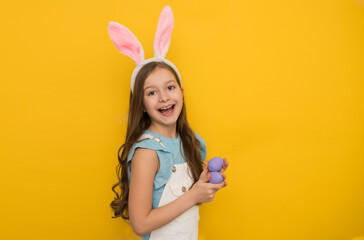 A cute girl with dark hair in a headband with rabbit ears holds purple Easter eggs in her hands and smiles without looking at the camera. Studio portrait on a yellow background. Happy childhood, happy