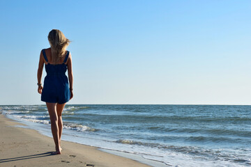 Fototapeta na wymiar A blonde girl in denim overalls is walking along the sea beach. Back view