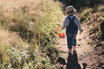 A little girl in a hat walks along the path on a clear, sunny summer day, holding a bucket of toys in her hands.