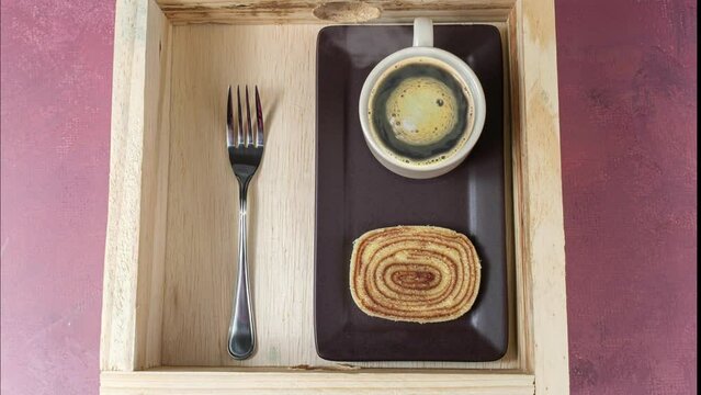 Slice of bolo de rolo next to cup of coffee and a fork over a wooden tray.
