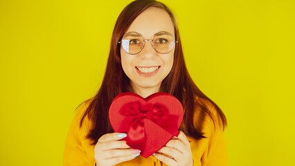 Young woman with red gift box in form of heart on yellow background. Pretty female holds romantic gift, looking at camera and smiling. Concept of present on valentine day and international womens day.